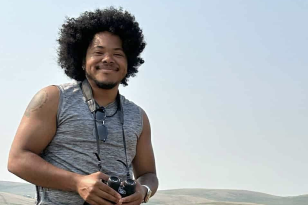 Smiling man with camera stands outdoors against a scenic backdrop, capturing the beauty of nature.